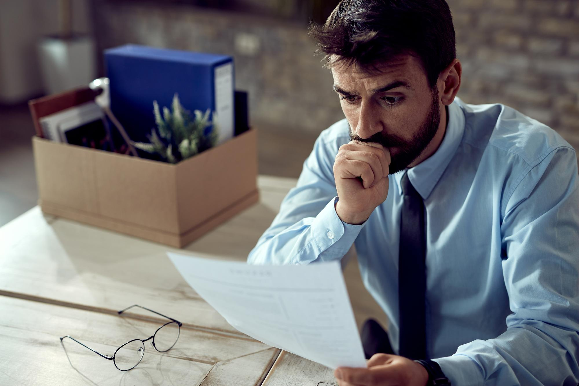 A imagem mostra um homem em um ambiente de escritório, sentado à mesa e analisando atentamente um documento impresso. Ele veste camisa social clara e gravata, transmitindo um contexto corporativo e profissional.