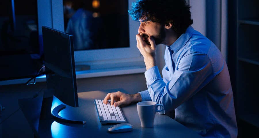 homem sentado em frente ao computador, dentro de um quarto, com a mão na boca e a outra no teclado