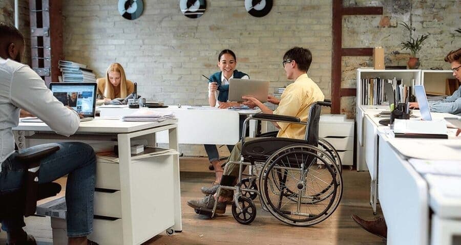 pessoas sentadas trabalhando numa mesa e um homem numa cadeira de rodas ao lado
