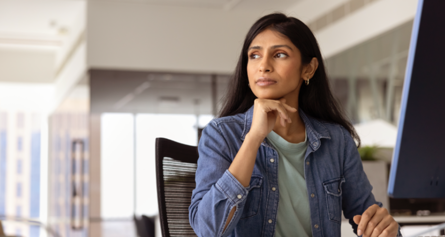 A imagem mostra uma mulher sentada em seu ambiente de trabalho, olhando para o lado com uma expressão pensativa, séria e um pouco desconfortável. Ela apoia o queixo na mão, como se estivesse refletindo profundamente ou tentando lidar com uma situação difícil.