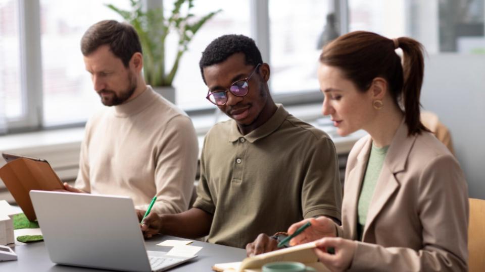 A imagem mostra três profissionais trabalhando juntos em um ambiente corporativo moderno e iluminado por luz natural. Eles estão sentados lado a lado em uma mesa de trabalho, utilizando notebooks, tablets e materiais de anotação durante uma reunião ou atividade colaborativa.