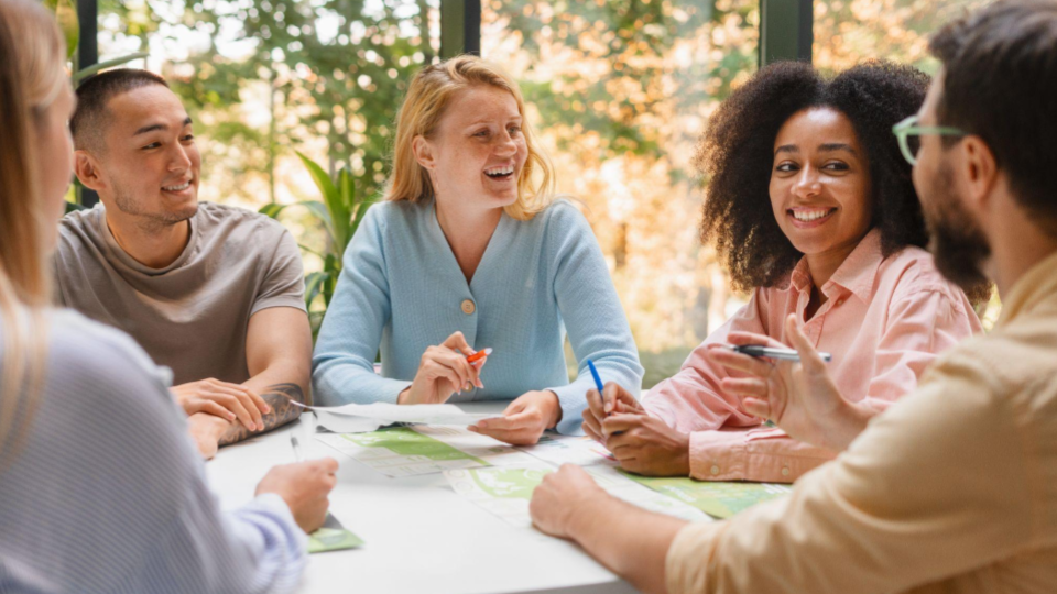 Esta imagem retrata uma reunião colaborativa e descontraída entre cinco jovens adultos em um ambiente bem iluminado. A atmosfera sugere um espaço de trabalho moderno ou uma sala de estudos com grandes janelas que mostram uma folhagem verde ao fundo.
