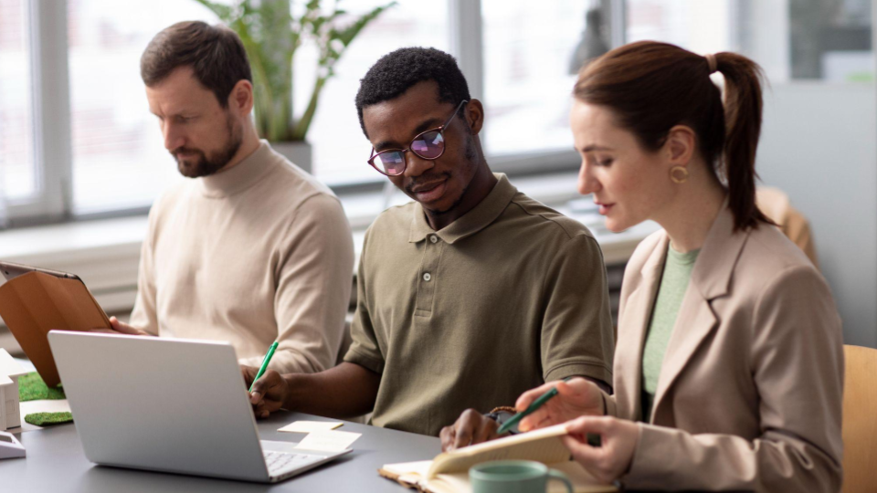 A imagem mostra três pessoas sentadas lado a lado em uma mesa de trabalho