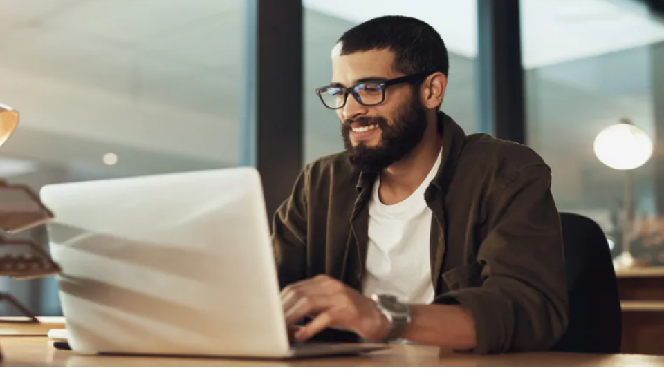 Um homem sorridente, com óculos e barba, está sentado à mesa trabalhando em um laptop em um ambiente de escritório moderno.
