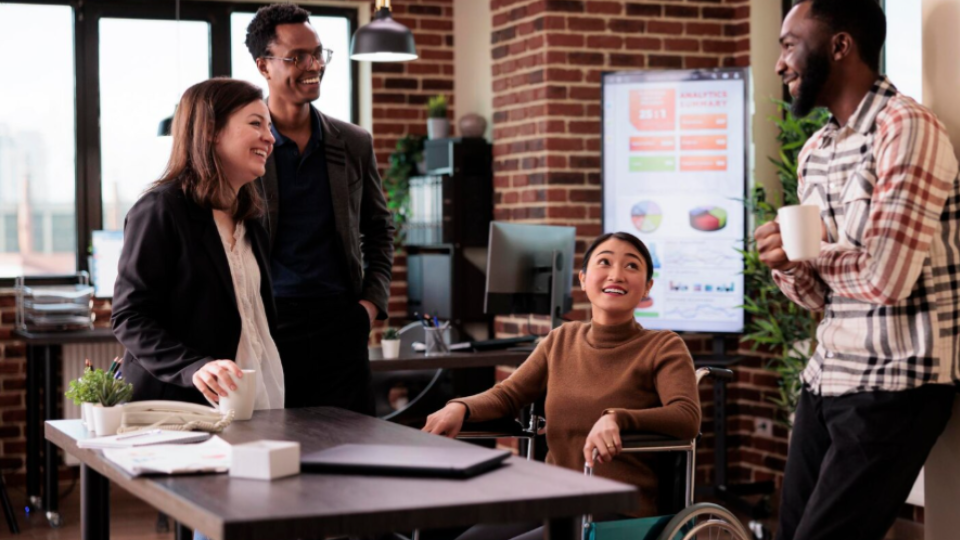 Equipe diversa reunida em um escritório moderno, conversando de forma colaborativa ao redor de uma mesa de trabalho. O ambiente é acolhedor e profissional, com destaque para a inclusão, representada por uma colaboradora em cadeira de rodas participando ativamente da interação. Ao fundo, há gráficos e indicadores exibidos em uma tela, sugerindo um contexto de negócios e planejamento estratégico.