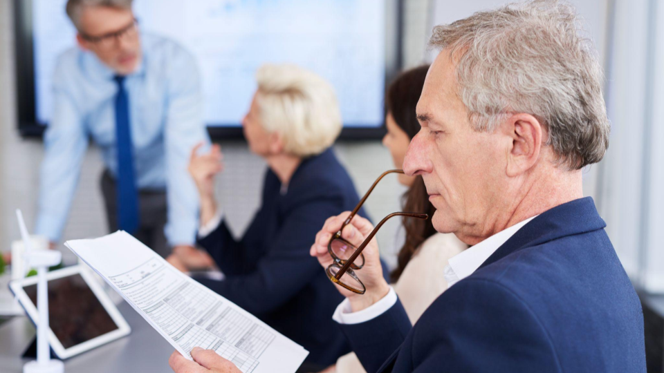 A imagem mostra uma reunião de negócios em um ambiente corporativo moderno, focando em executivos analisando dados e relatórios.