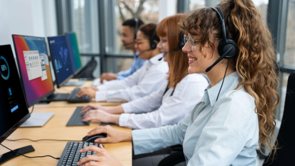 Fotografia realista de uma equipe de atendimento ao cliente trabalhando em um ambiente corporativo moderno. Os profissionais estão sentados lado a lado, utilizando computadores, teclados e headsets com microfone para realizar atendimentos.