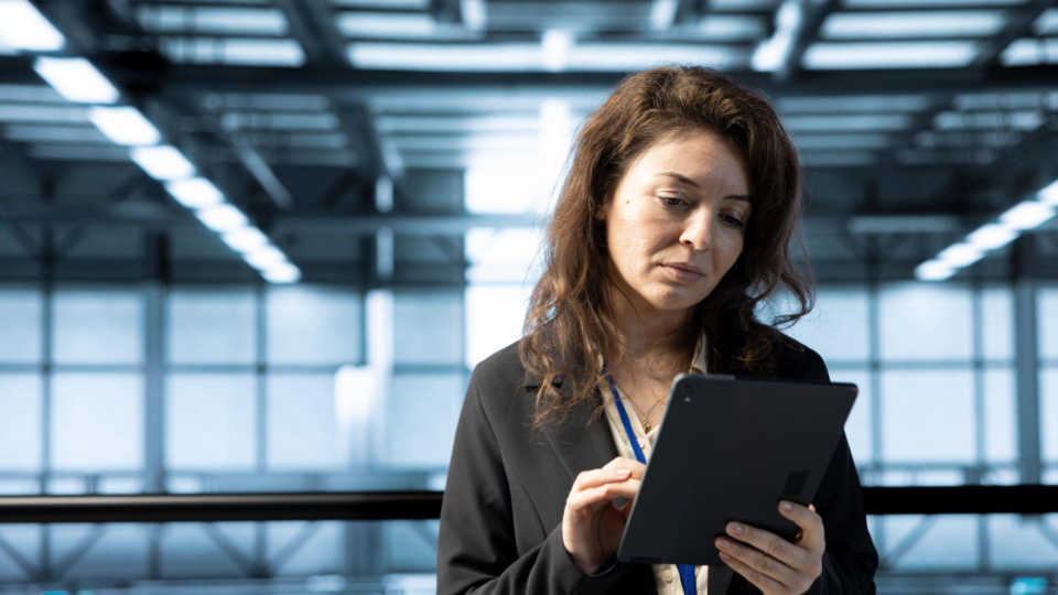 Esta imagem mostra uma mulher em um ambiente corporativo ou industrial moderno, concentrada ao utilizar um dispositivo móvel.