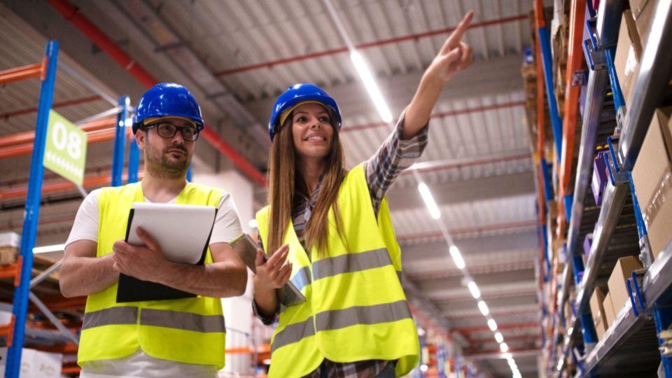 Dois profissionais de logística usando capacetes e coletes de segurança enquanto inspecionam o estoque em um centro de distribuição, representando a segurança e o controle na cadeia de suprimentos.