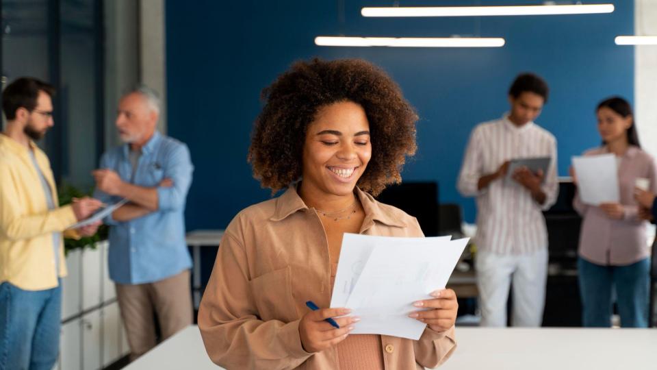 A imagem mostra um ambiente corporativo moderno e colaborativo, focado em uma mulher em primeiro plano com outros colegas trabalhando ao fundo.