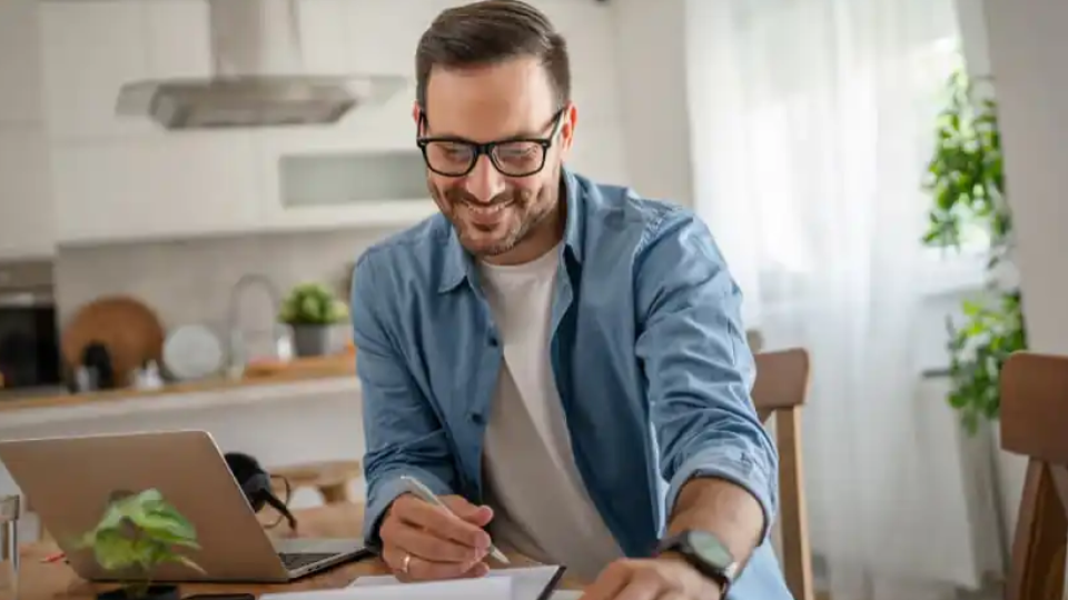 Um homem sorridente trabalha em casa, sentado à mesa da cozinha em um ambiente moderno e iluminado.