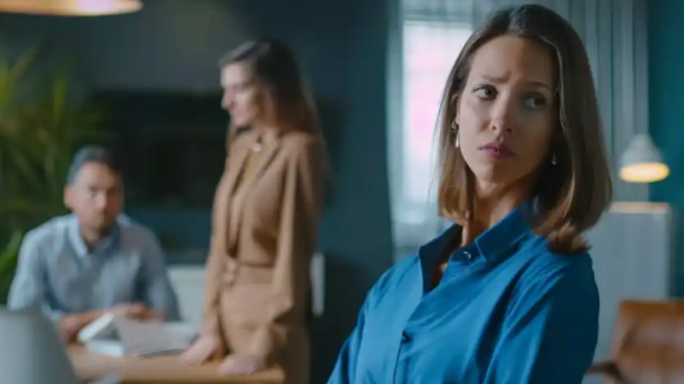 A imagem mostra um ambiente corporativo com clima de tensão ou preocupação. Em primeiro plano, uma mulher usando camisa azul aparece em destaque, olhando para o lado com expressão séria e reflexiva.
