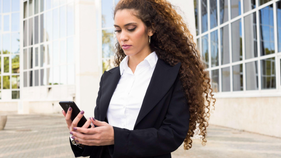 Uma mulher de negócios com longos cabelos cacheados e volumosos está em pé, segurando e olhando atentamente para um smartphone. Ela veste um traje formal composto por um blazer preto sobre uma camisa social branca, transmitindo uma imagem de seriedade e profissionalismo.