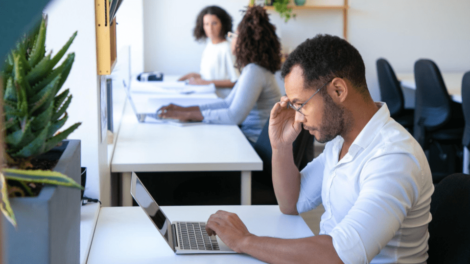 A imagem mostra um ambiente de escritório moderno e bem iluminado, com três pessoas trabalhando em computadores ao longo de uma bancada branca próxima a uma janela.
