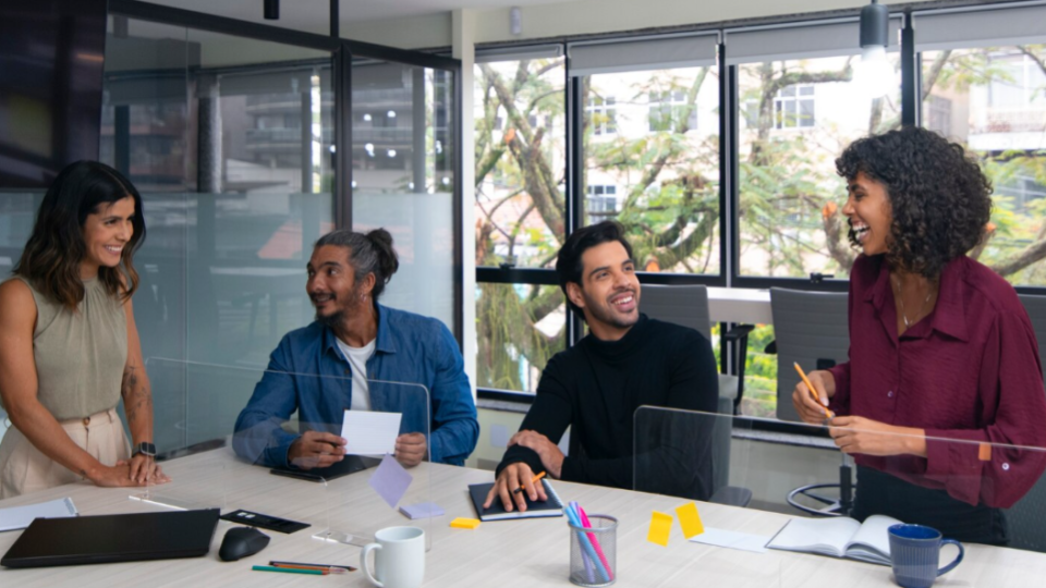 A imagem mostra um ambiente de escritório moderno e colaborativo, com quatro pessoas reunidas em torno de uma mesa de conferência clara. O clima geral é descontraído, profissional e amigável.