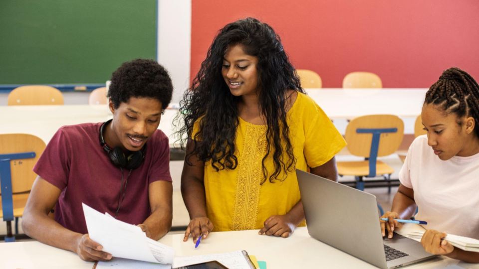 Três jovens adultos em uma sala de aula colaborando sobre documentos e utilizando um notebook, representando o ambiente seguro e transparente promovido por um canal de denúncias em escolas e ONGs.
