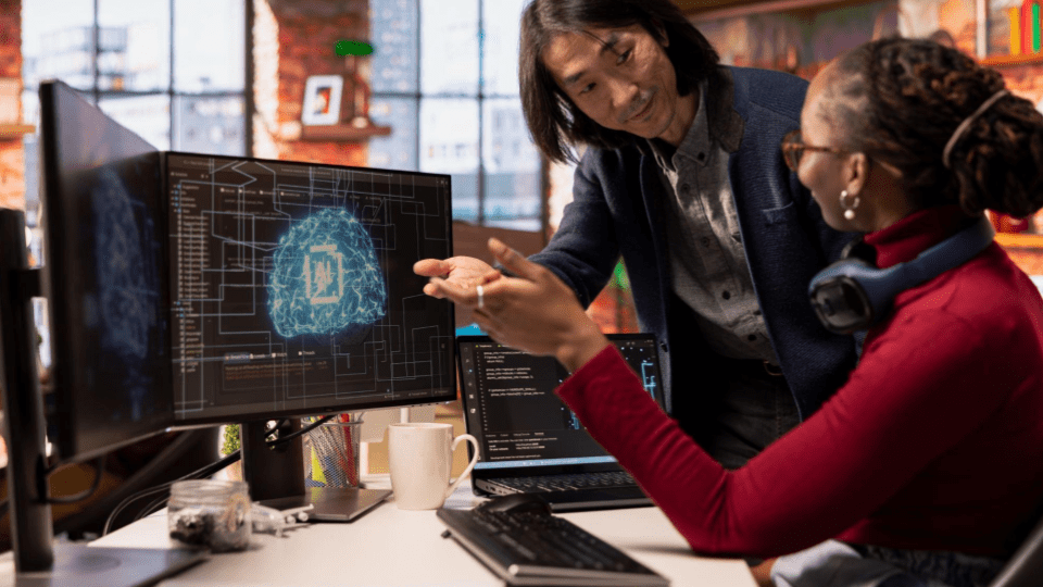 A imagem retrata um ambiente de trabalho moderno e colaborativo, focado em tecnologia de ponta. Nela, vemos dois profissionais — um homem e uma mulher — analisando dados em um escritório com estética industrial (tijolos aparentes ao fundo).