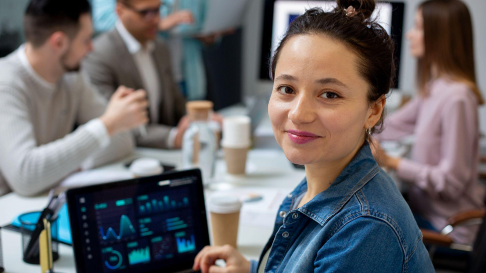 Uma jovem mulher em destaque, sorrindo para a câmera. Ela tem o cabelo preso em um coque alto e veste uma jaqueta jeans azul. Sua expressão transmite confiança, simpatia e engajamento.