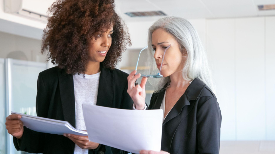 A imagem mostra duas mulheres em um ambiente corporativo moderno, aparentemente dentro de um escritório bem iluminado.