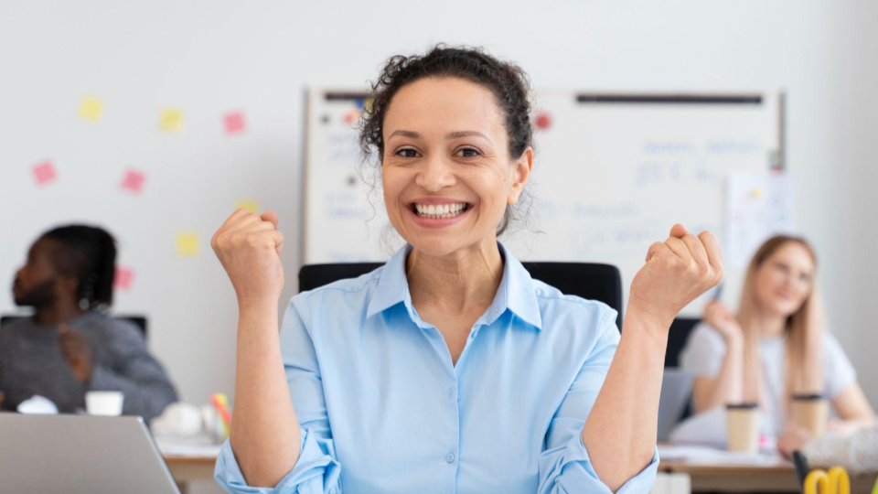 A imagem mostra uma mulher sorridente num ambiente moderno de escritório, celebrando uma conquista ou sucesso.