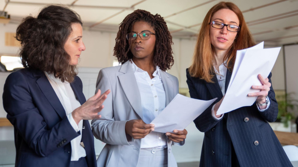 Esta imagem retrata três mulheres em trajes corporativos formais, aparentemente engajadas em uma reunião de negócios ou discussão de trabalho em um ambiente de escritório.
