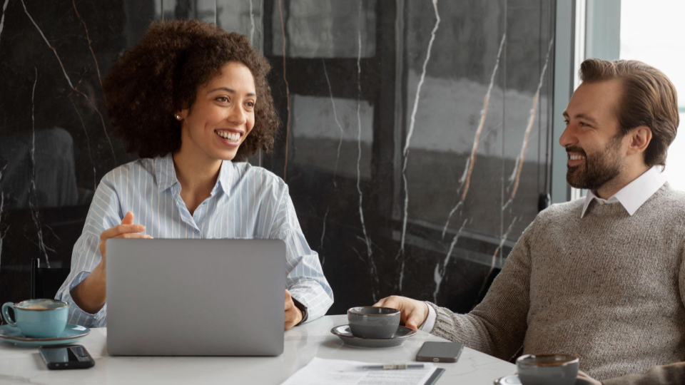 A imagem mostra dois profissionais em uma reunião informal em um ambiente corporativo moderno. Sentados à mesa, eles conversam de forma descontraída, sorrindo e mantendo contato visual, o que transmite um clima de colaboração, confiança e engajamento.