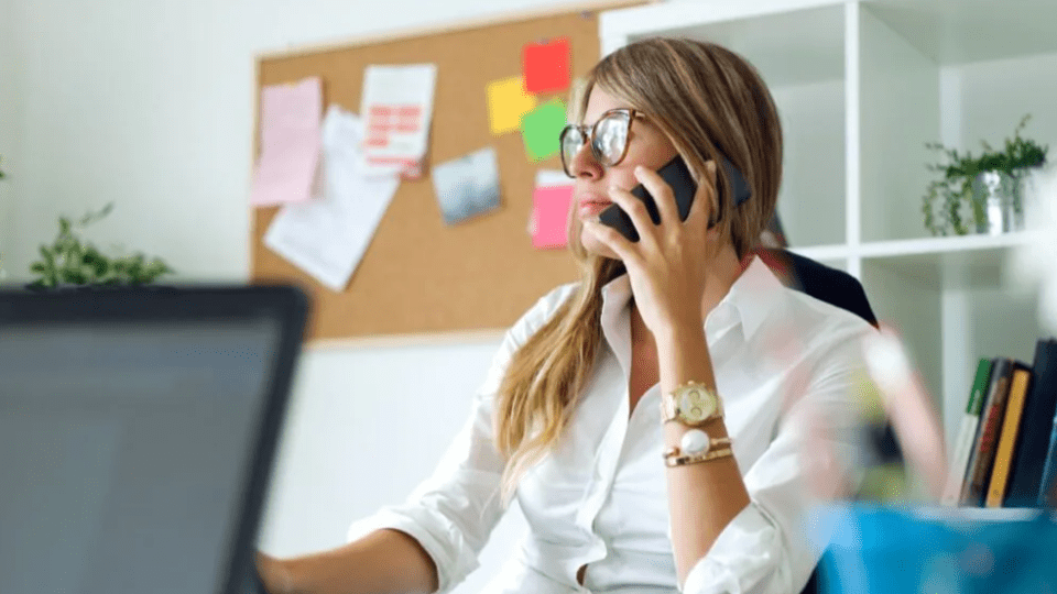 A imagem mostra uma mulher em um ambiente de escritório, sentada à mesa enquanto fala ao telefone celular.