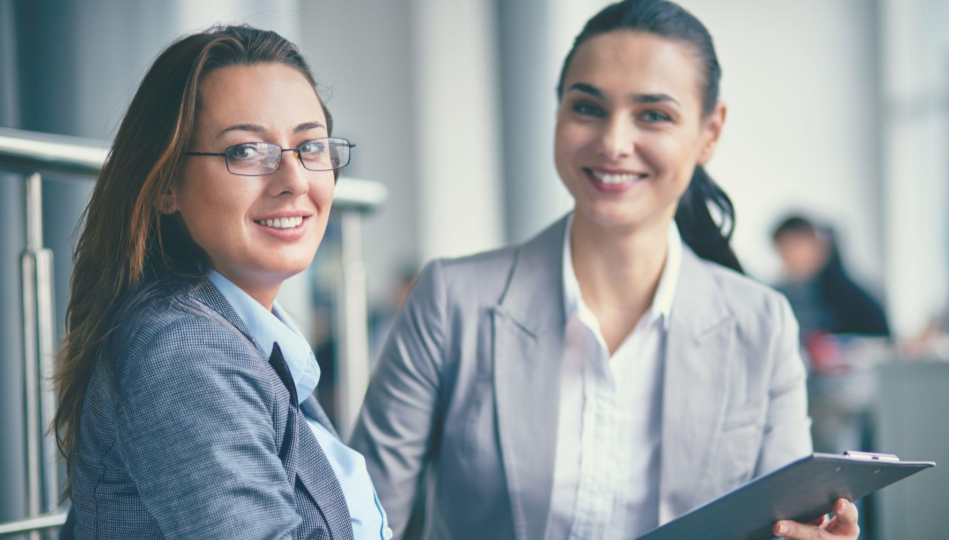 A imagem retrata duas mulheres em um ambiente corporativo moderno, transmitindo profissionalismo e simpatia.