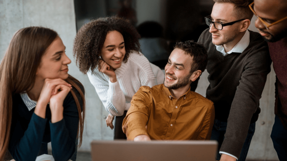 A imagem mostra um grupo de cinco pessoas em um ambiente de trabalho colaborativo, reunidas em torno de um notebook. Todos parecem jovens adultos e estão próximos uns dos outros, em uma interação descontraída e positiva.