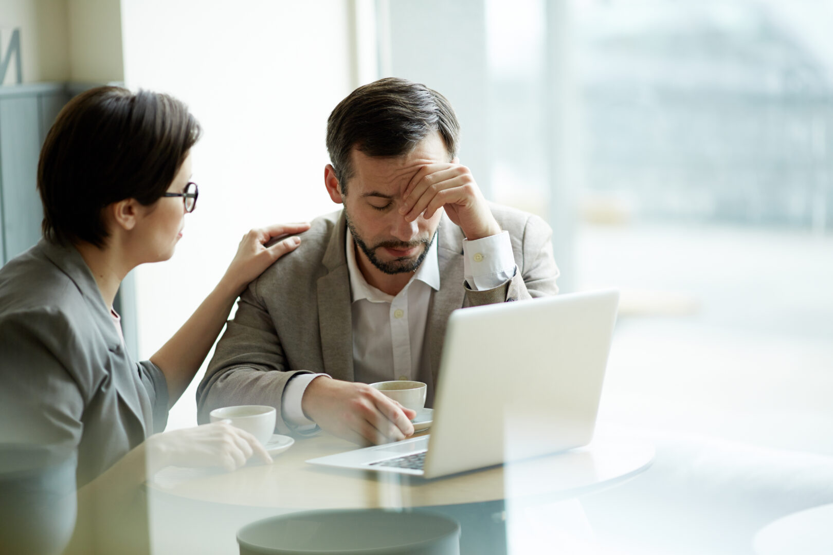 Na imagem, há duas pessoas em um ambiente corporativo, sentadas à mesa com um notebook aberto e xícaras de café.