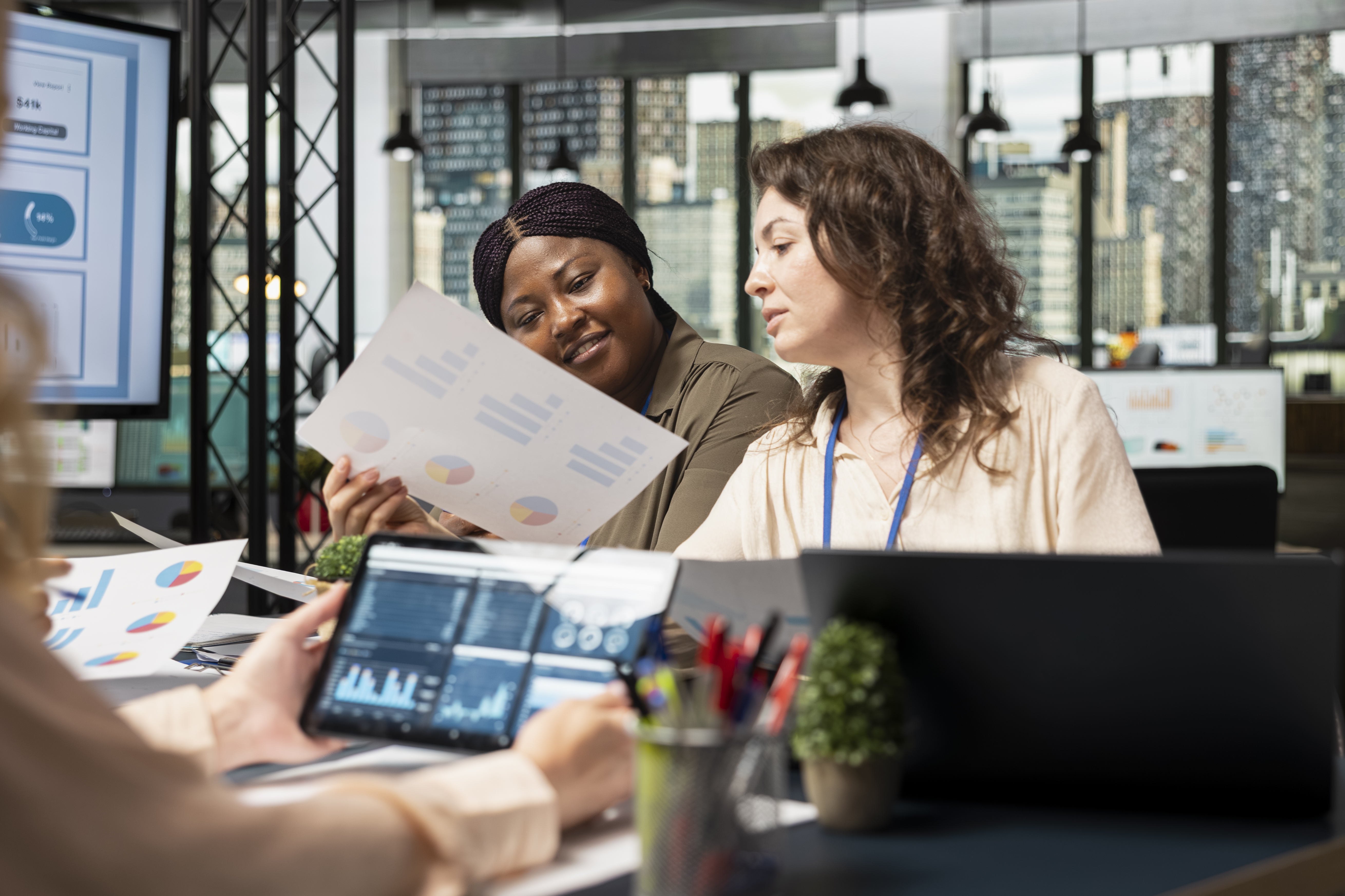 A imagem mostra uma mulher sentada à mesa de trabalho em um ambiente de escritório, aparentando preocupação ou cansaço. Ela apoia a cabeça com uma das mãos enquanto analisa papéis e contas, olhando atentamente para os documentos.
