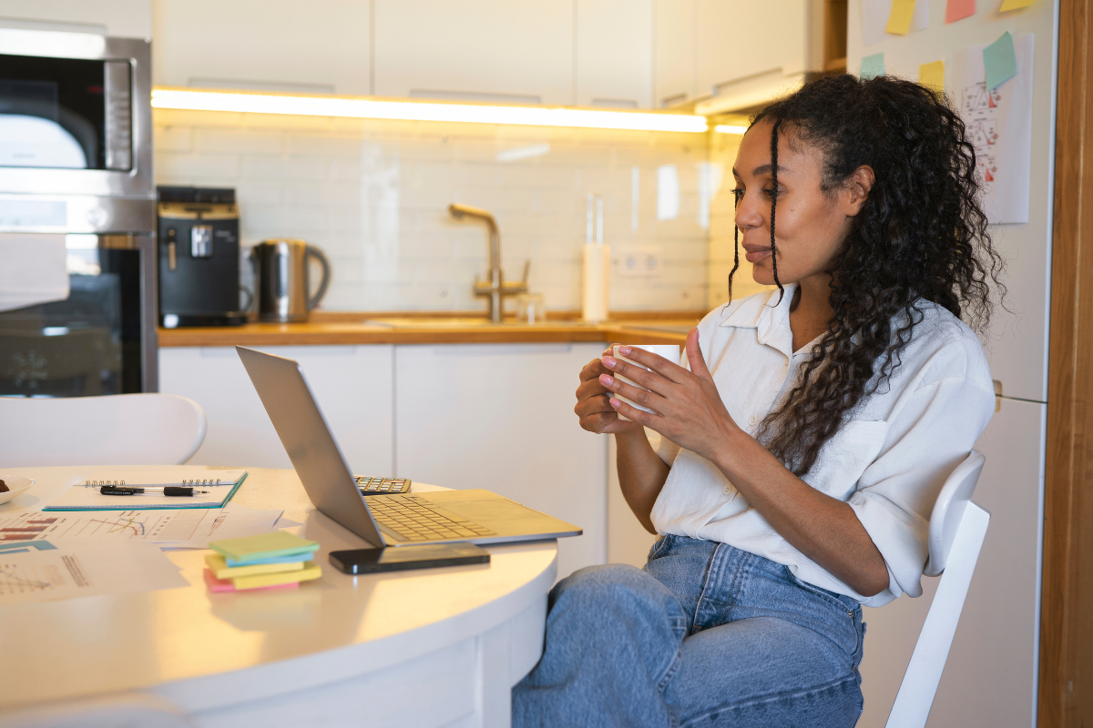 A imagem mostra uma mulher trabalhando em casa, sentada à mesa de uma cozinha moderna e bem iluminada. Ela está usando um notebook aberto à sua frente e segura uma xícara com as duas mãos, como se estivesse fazendo uma pausa para café enquanto analisa algo na tela.